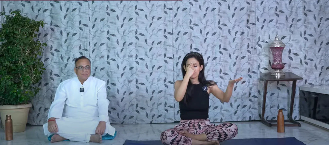 Two people seated cross-legged on yoga mats indoors, practicing a guided breathing exercise, with one person demonstrating hand movements for pranayama in a calm wellness setting.
